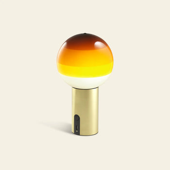 The Marset Dipping Light Portable by Marset features a cylindrical gold base and a colored glass sphere shade with gradient hues from dark brown to orange, yellow, and white, creating a striking ambient effect against any beige background.