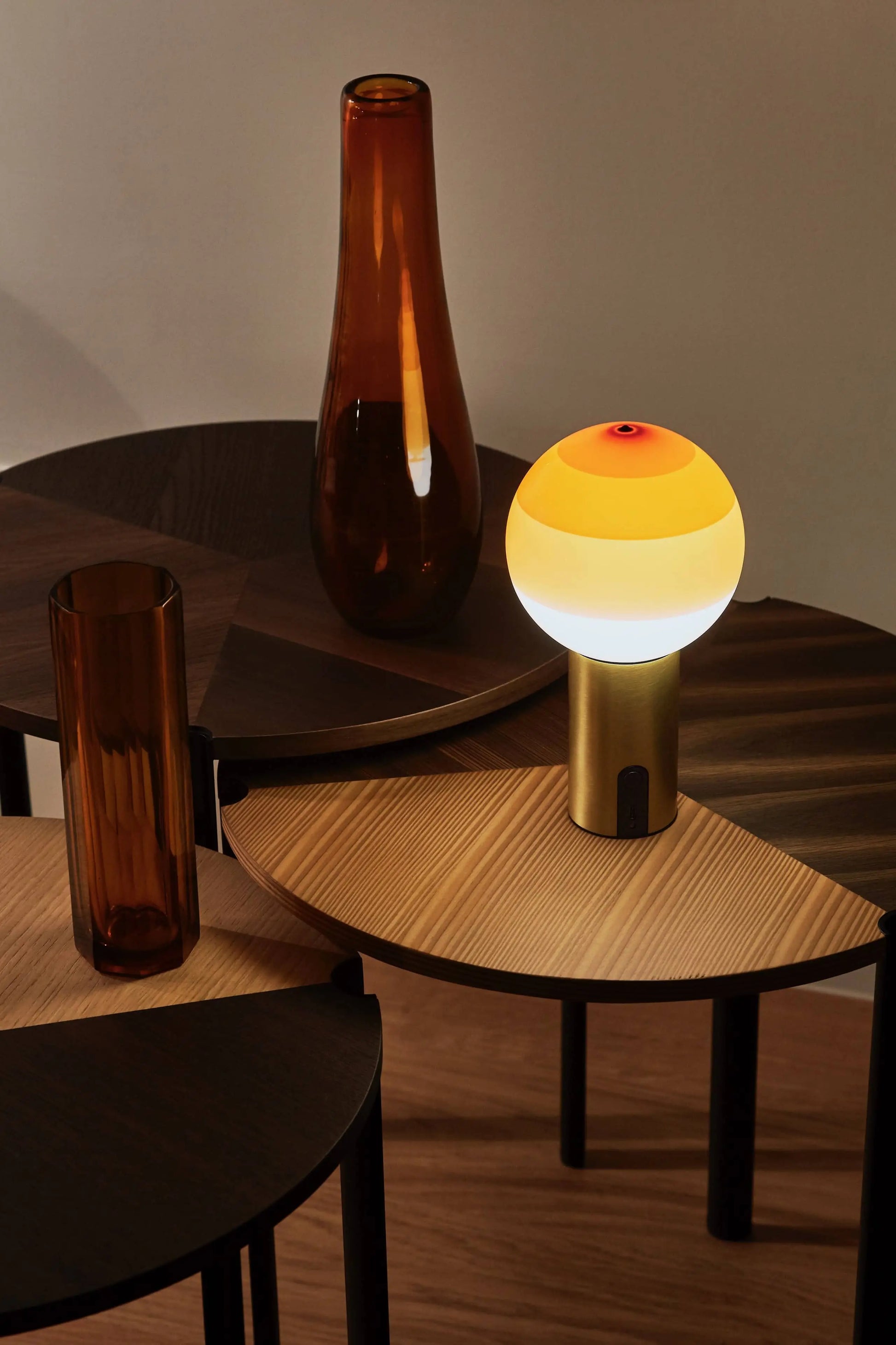 Three round wooden side tables with two amber glass vases and the Marset Dipping Light Portable by Marset—a colored glass sphere lamp with a glowing yellow and white shade—create a warm, minimalist, and inviting ambience.