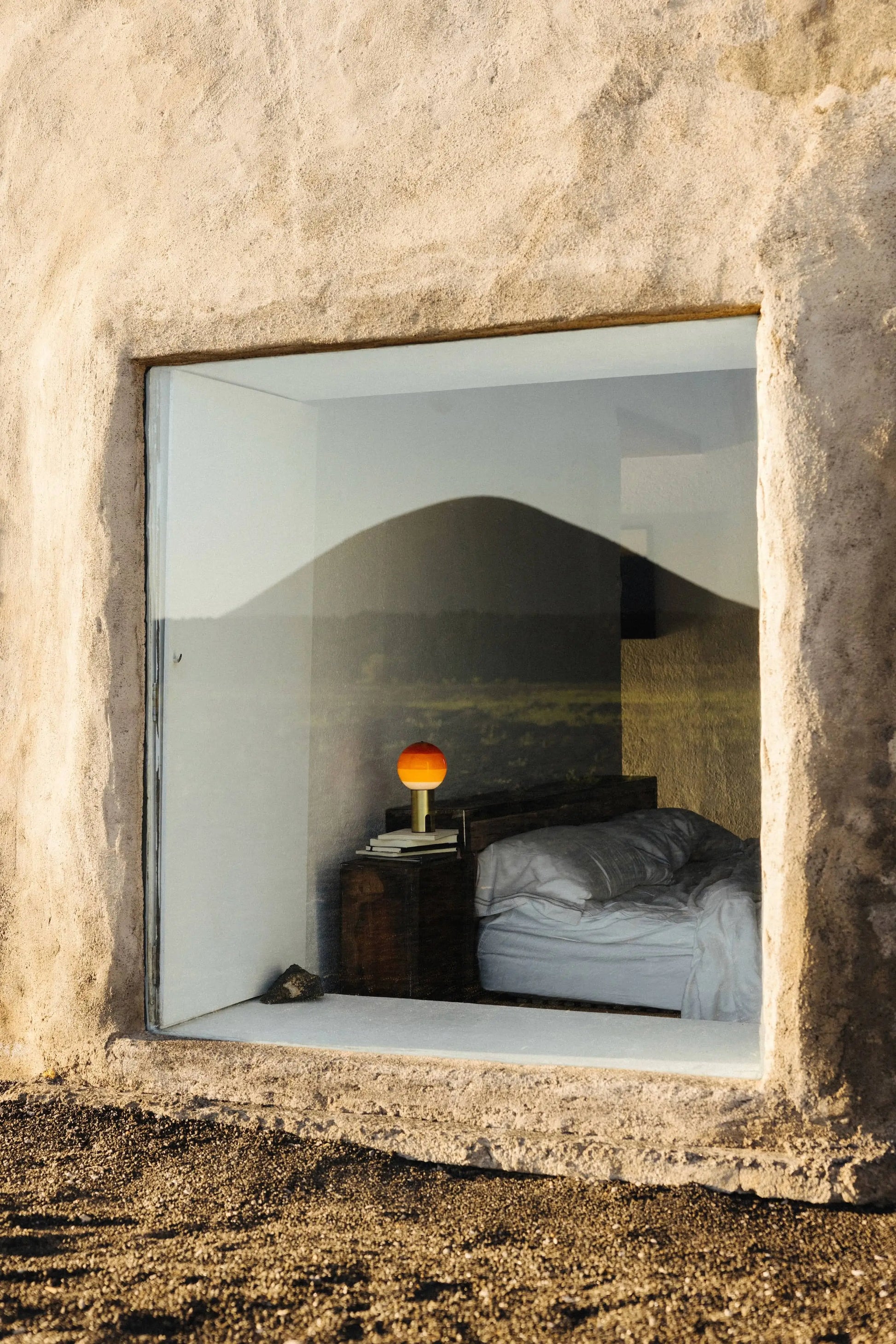 A large window reveals a cozy bedroom with an unmade bed, wooden nightstand, stack of books, and the Marset Dipping Light Portable by Marset. The window reflects mountains and a landscape, creating an ambient glow in the room.