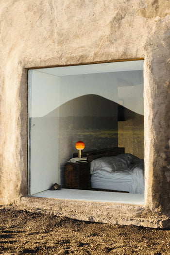 A large window reveals a cozy bedroom with an unmade bed, wooden nightstand, stack of books, and the Marset Dipping Light Portable by Marset. The window reflects mountains and a landscape, creating an ambient glow in the room.