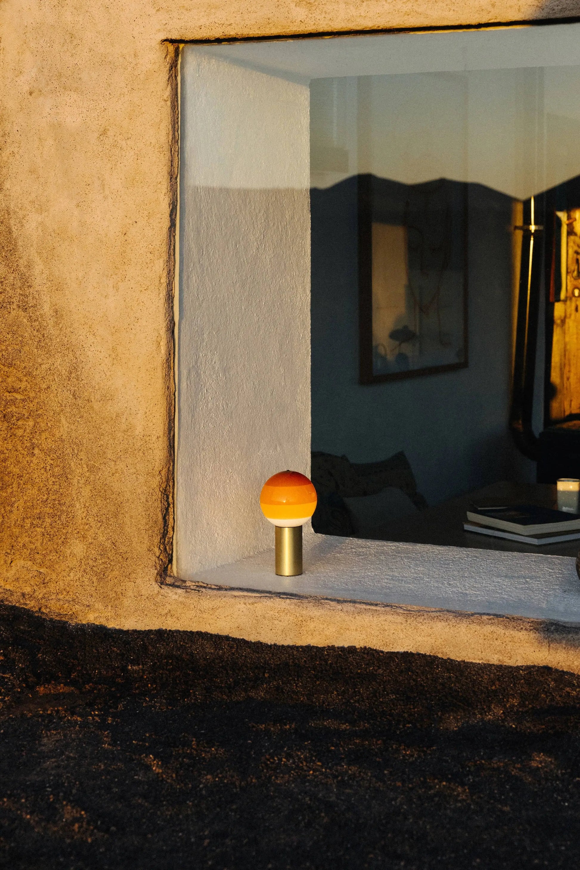A Marset Dipping Light Portable with an orange dome rests on a windowsill, glowing warmly in sunlight. The coloured glass sphere by Marset casts ambient light, while the window shows cozy, art-filled interiors and outdoor scenery.