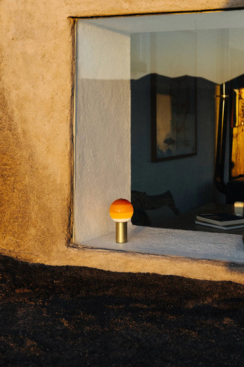 A Marset Dipping Light Portable with an orange dome rests on a windowsill, glowing warmly in sunlight. The coloured glass sphere by Marset casts ambient light, while the window shows cozy, art-filled interiors and outdoor scenery.