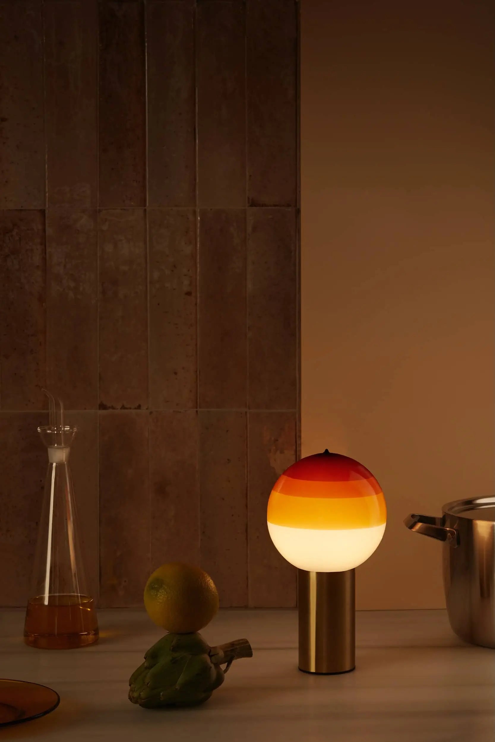 A modern table setting features the Marset Dipping Light Portable by Marset with a coloured glass sphere, a glass carafe with amber liquid, stainless steel pot, orange, and limes on marble against a brown tiled wall for an inviting ambiance.