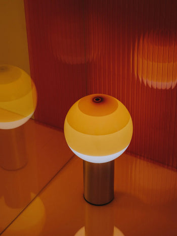 The Marset Dipping Light Portable by Marset, with its round coloured glass shade and metallic base, sits on a glossy surface, casting a warm ambient glow and reflections against an orange textured wall.