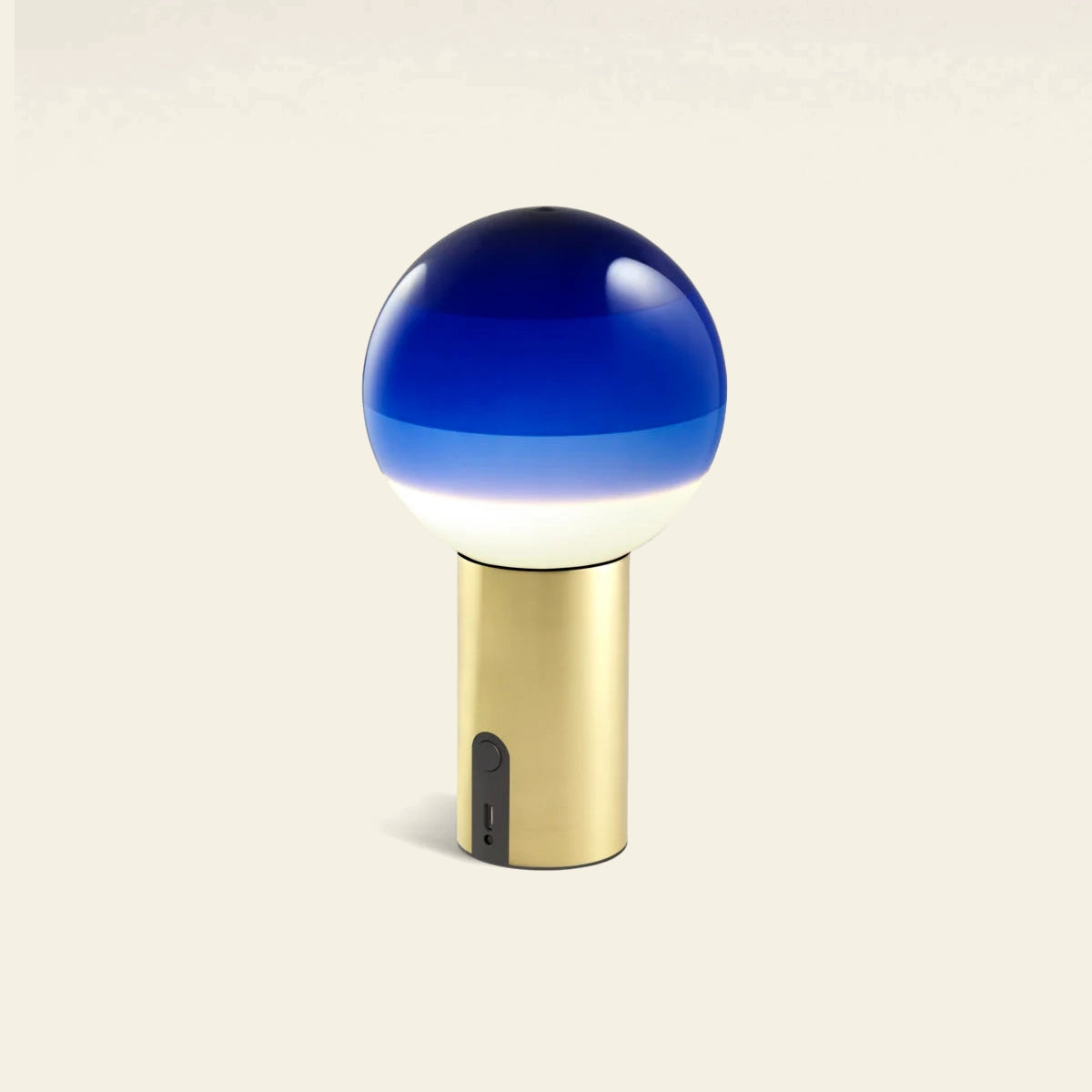 The Marset Dipping Light Portable by Marset features a blue and white gradient glass sphere atop a gold cylindrical base with an on/off switch and USB port, creating ambient light against a plain background.