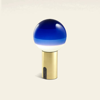 The Marset Dipping Light Portable by Marset features a blue and white gradient glass sphere atop a gold cylindrical base with an on/off switch and USB port, creating ambient light against a plain background.