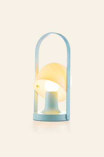 The Marset FollowMe Portable Table Lamp by Marset features a pastel blue base, arched handle, and tilted yellow shade, casting soft light on a plain beige background.
