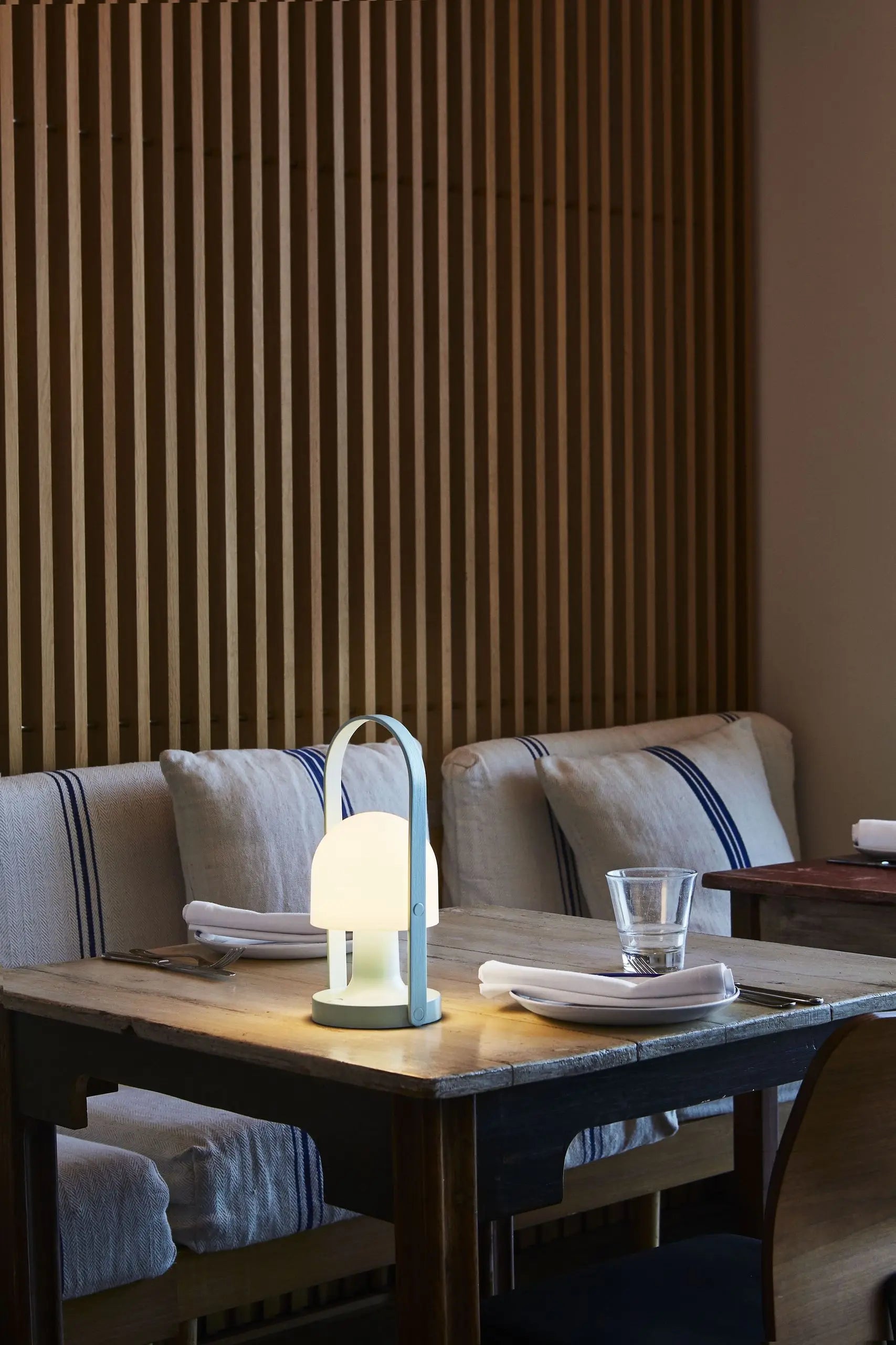 A cozy restaurant table for two features the Marset FollowMe Portable Table Lamp, a glass, and neatly folded napkins. Cushioned benches with striped pillows sit against a wooden slat wall.