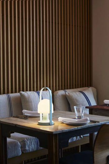 A cozy restaurant table for two features the Marset FollowMe Portable Table Lamp, a glass, and neatly folded napkins. Cushioned benches with striped pillows sit against a wooden slat wall.