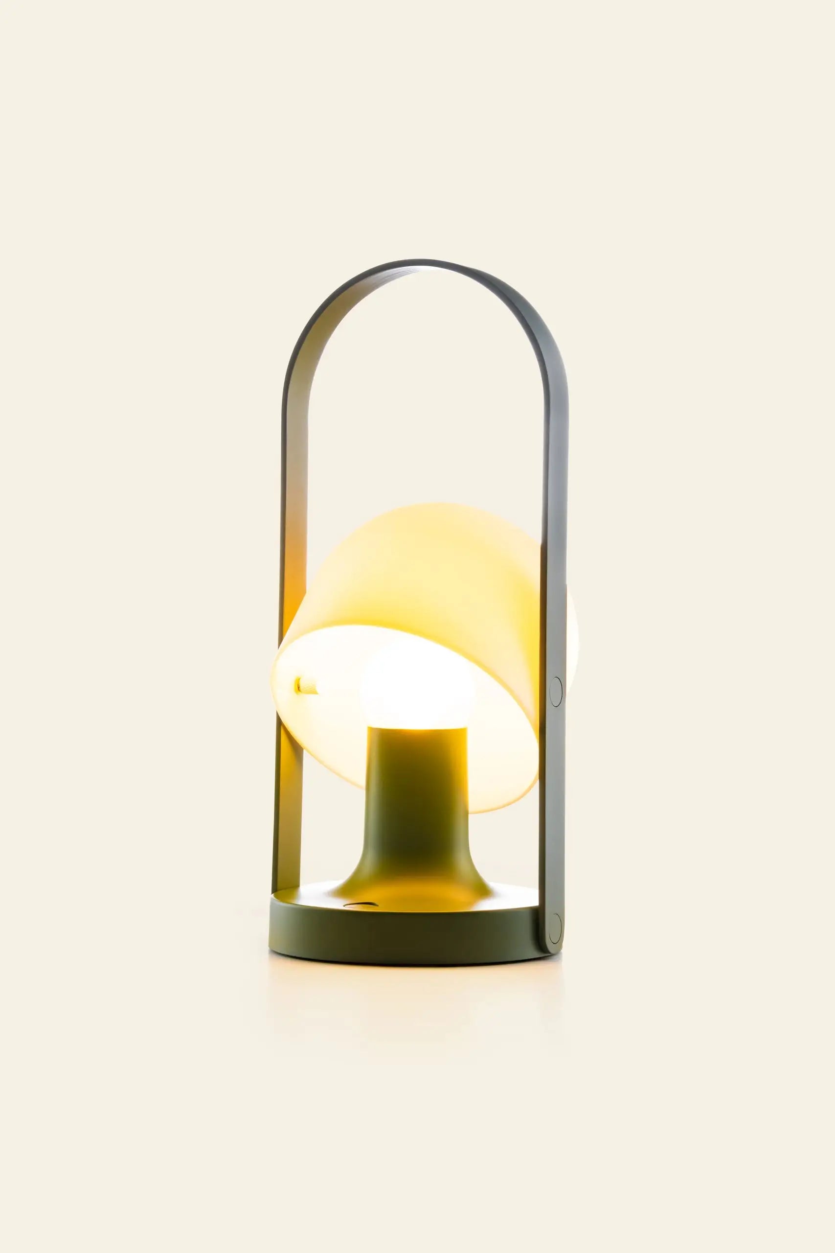 The Marset FollowMe Portable Table Lamp by Marset features a modern design with a round yellow-tinged shade, dark gray handle, and rechargeable battery, casting a warm light against a plain cream background.