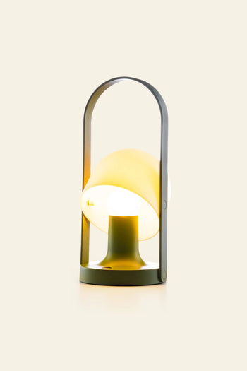 The Marset FollowMe Portable Table Lamp by Marset features a modern design with a round yellow-tinged shade, dark gray handle, and rechargeable battery, casting a warm light against a plain cream background.