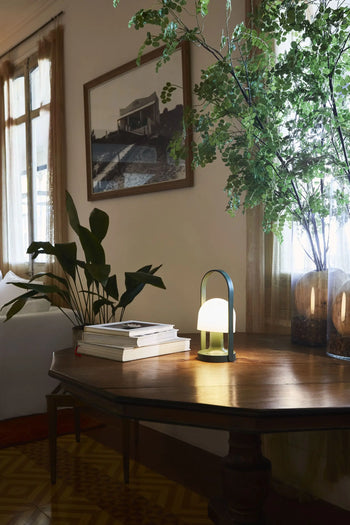 A wooden table with stacked books, the Marset FollowMe Portable Table Lamp by Marset with an oak handle, and vases of leafy branches sits beside a window with sheer curtains in a cozy, softly-lit living room. A framed photo hangs above.