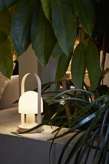 The Marset FollowMe Portable Table Lamp by Marset features a modern dome shape, oak handle, and rechargeable design. It emits a warm light, shown on a white ledge amid lush green leaves and indoor plants.