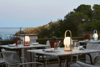 Outdoor restaurant tables with modern lanterns, glasses, napkins, and potted plants overlook the sea and rocky coastline. Marset FollowMe Portable Table Lamps by Marset, with oak handles, add elegance beneath trees and a soft evening sky.