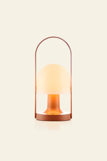 The Marset FollowMe Portable Table Lamp by Marset features a frosted dome-shaped shade, warm orange base, and curved oak handle. This modern, rechargeable lamp is shown against a plain beige background.