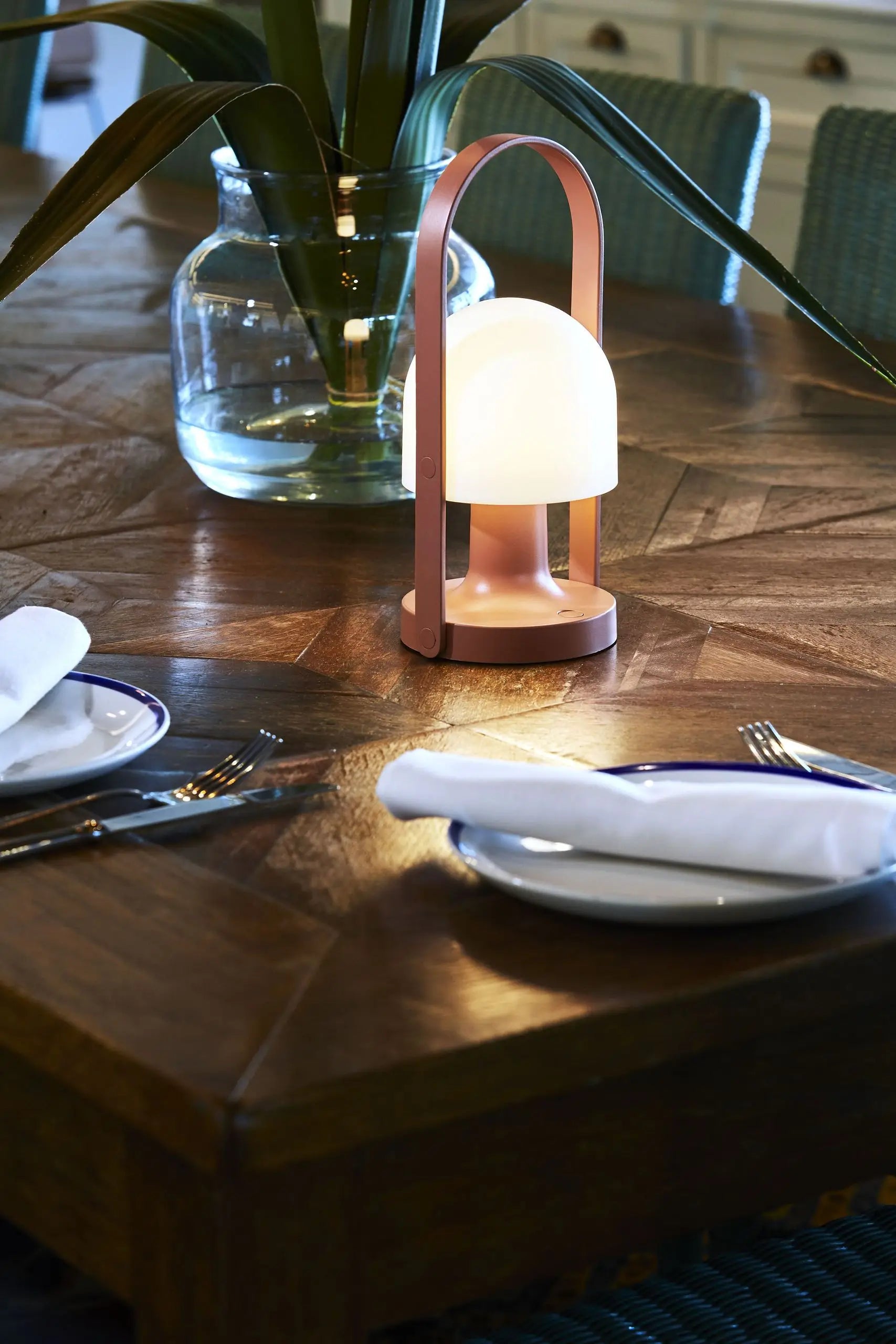 The Marset FollowMe Portable Table Lamp, a modern lamp with a dome-shaped shade and oak handle by Marset, sits on a wooden dining table set for two; in the background is a glass vase with green leaves.