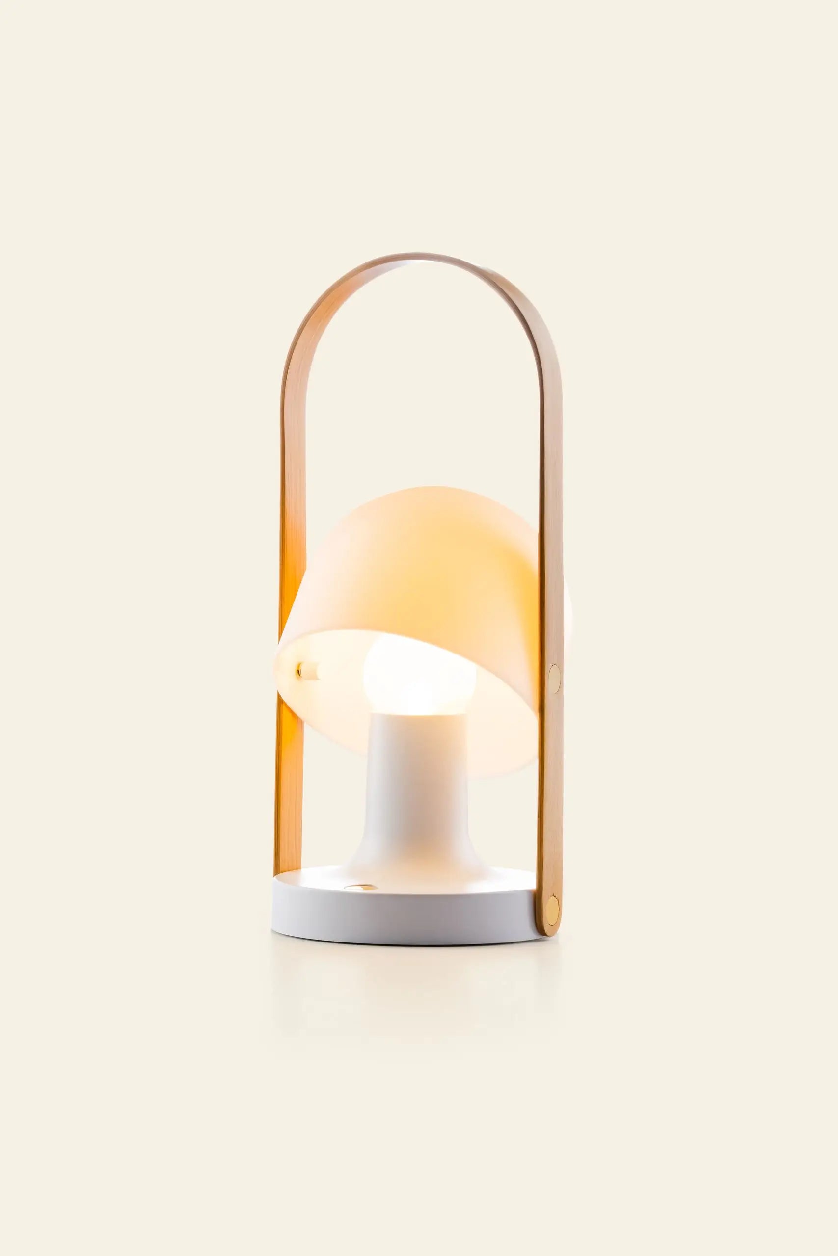 The Marset FollowMe Portable Table Lamp by Marset features a white round base, a tilted cream lampshade, and an oak handle gracefully arching over the top, shown against a plain beige background.