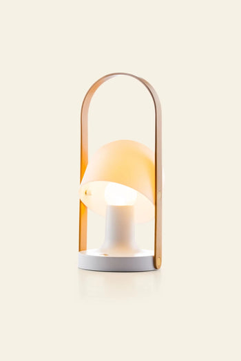 The Marset FollowMe Portable Table Lamp by Marset features a white round base, a tilted cream lampshade, and an oak handle gracefully arching over the top, shown against a plain beige background.