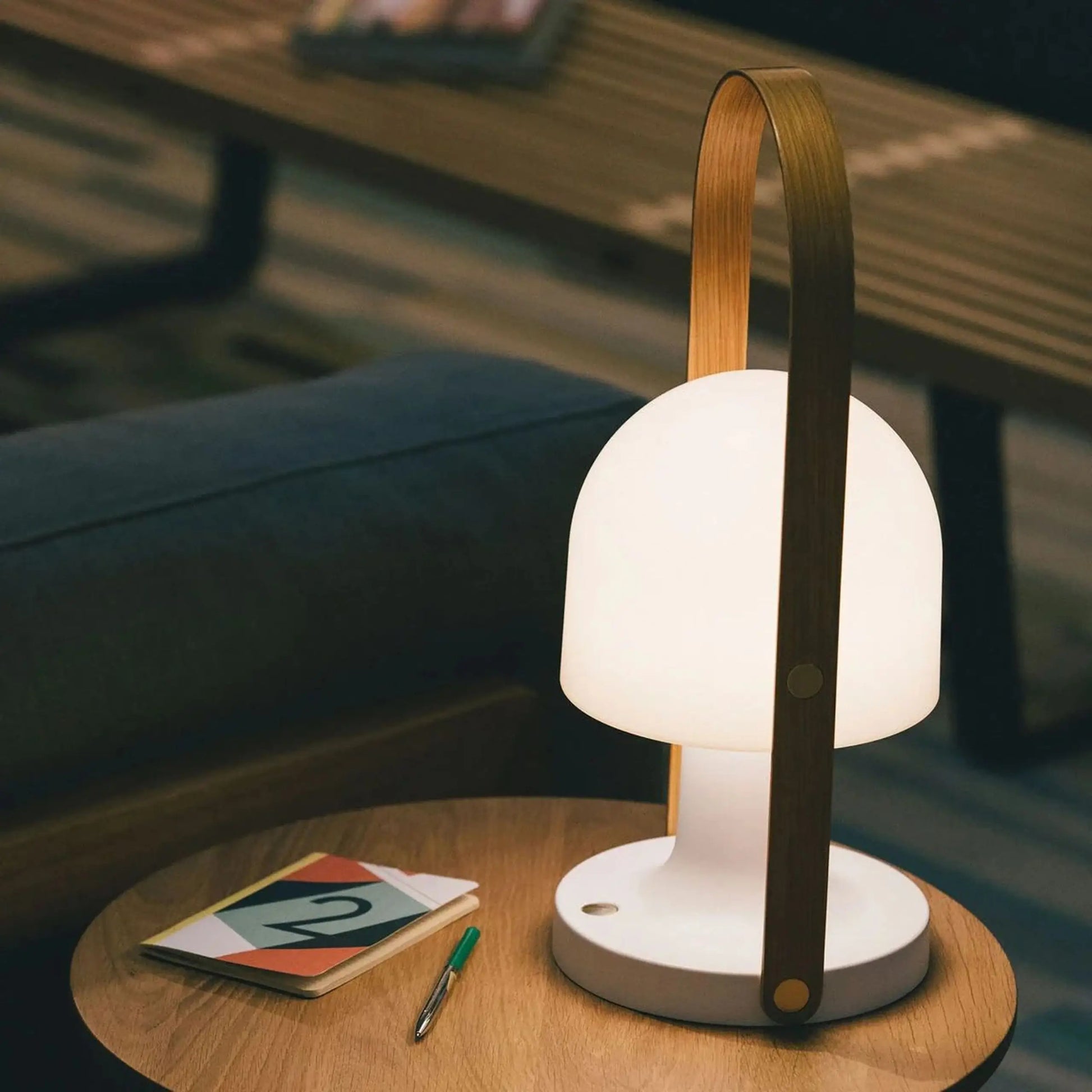 The Marset FollowMe Portable Table Lamp by Marset, featuring a modern white design and oak handle, sits on a round wooden table beside a colorful notepad, pen, and sofa. The rechargeable lamp is on, casting a warm glow.
