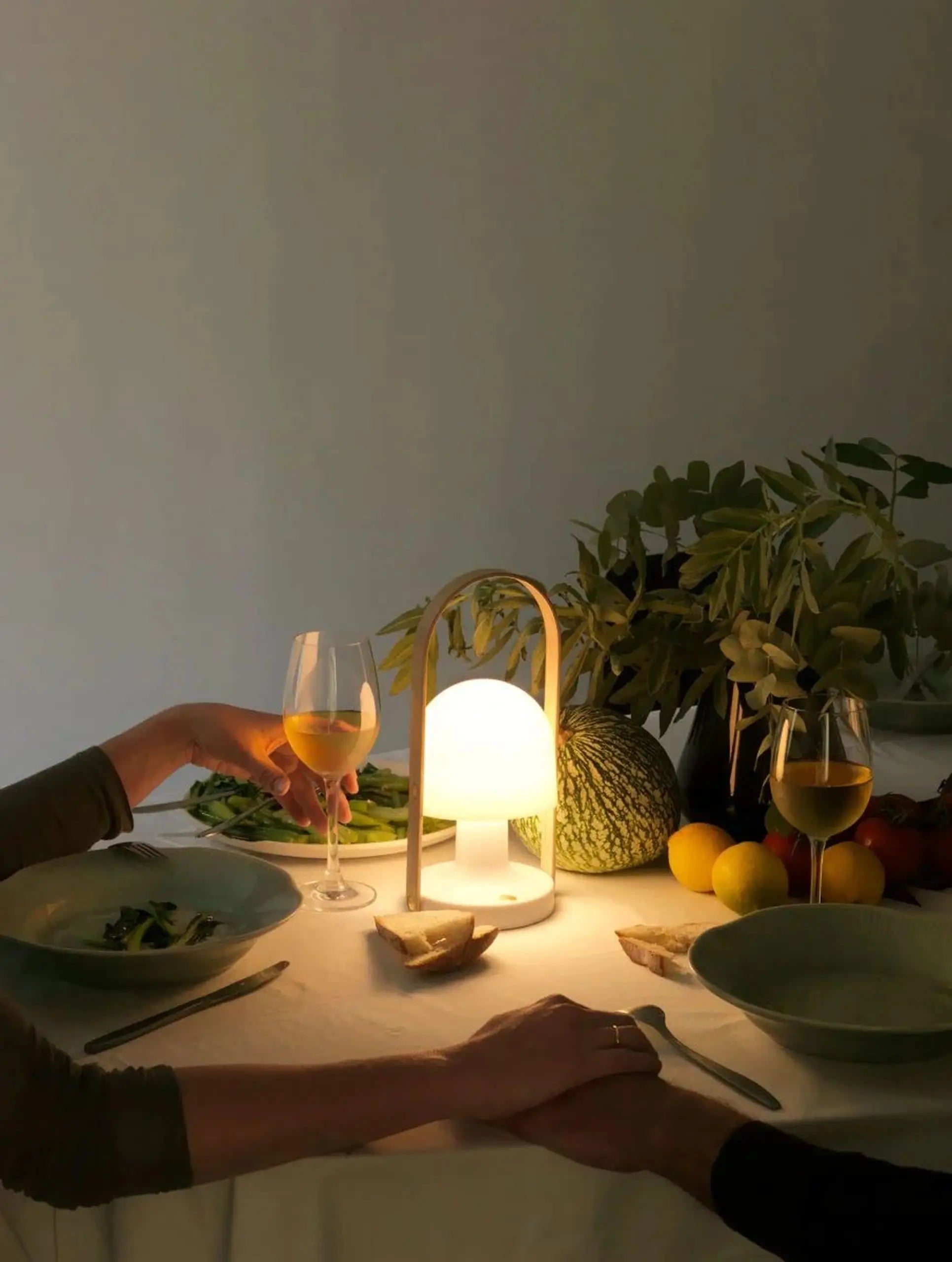 A softly lit dinner table features two people holding hands, salad plates, white wine glasses, and a glowing Marset FollowMe Portable Table Lamp by Marset, surrounded by fruits and greenery for an intimate, cozy atmosphere.