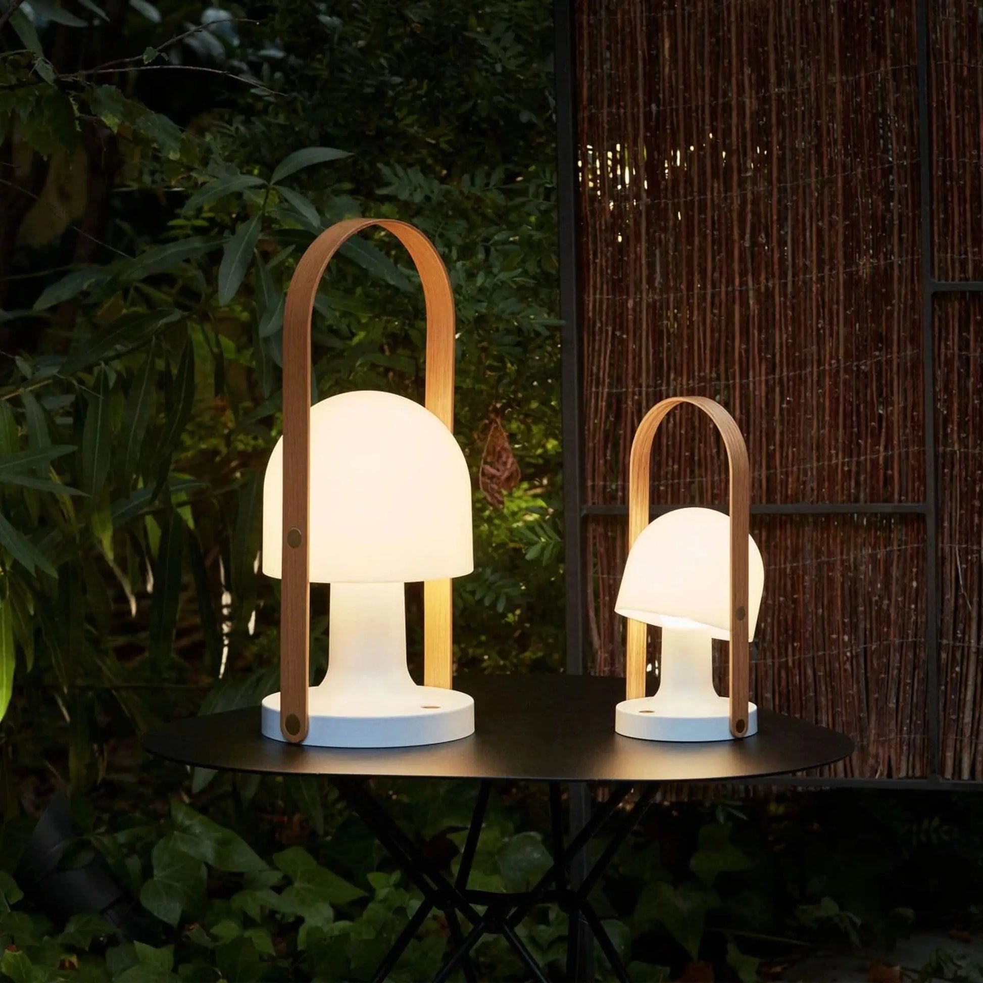 Two Marset FollowMe Portable Table Lamps, featuring oak handles and glowing white shades, sit on a round black outdoor table surrounded by greenery and a bamboo privacy screen in a nighttime garden.
