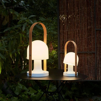 Two Marset FollowMe Portable Table Lamps, featuring oak handles and glowing white shades, sit on a round black outdoor table surrounded by greenery and a bamboo privacy screen in a nighttime garden.