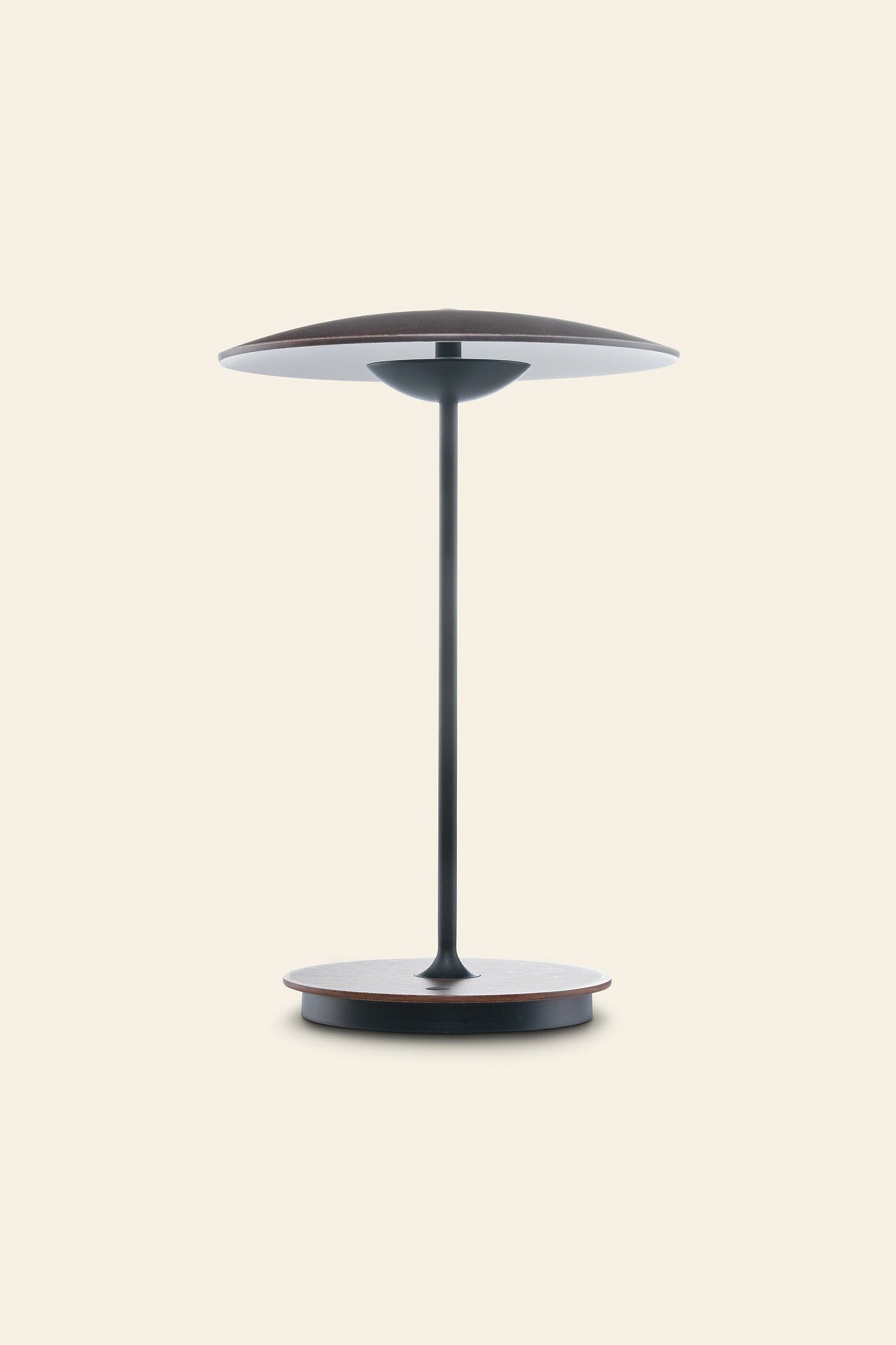 The Marset Ginger 20 M Table Lamp by Marset features a circular base, disc-shaped shade in a dark finish, and slim stem—ideal for modern decor and creating a cozy atmosphere against a plain light background.