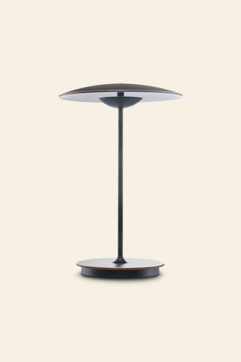 The Marset Ginger 20 M Table Lamp by Marset features a circular base, disc-shaped shade in a dark finish, and slim stem—ideal for modern decor and creating a cozy atmosphere against a plain light background.