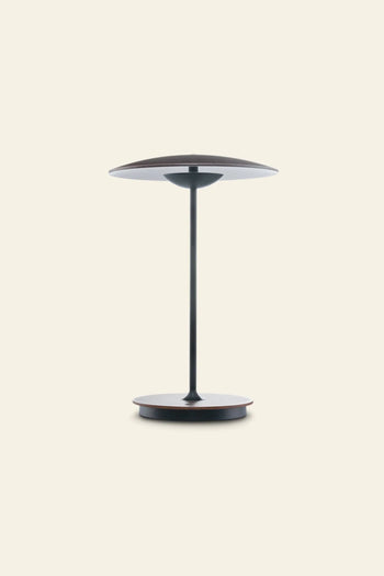 The Marset Ginger 20 M Table Lamp by Marset features a minimalist round design with a slim black metal base and double wooden shades, ideal for creating a cosy modern atmosphere against a plain beige backdrop.