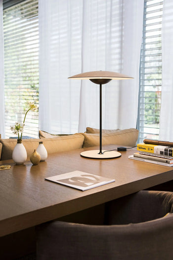 A modern desk featuring the Marset Ginger 20 M Table Lamp by Marset, stacked books, vases, and a magazine sits before a window with blinds and sheer curtains. Cushions line the seat, adding coziness to the contemporary decor.