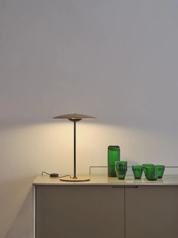 The Marset Ginger 20 M Table Lamp from Marset casts a warm glow over a light gray cabinet, highlighting green glass vases and tumblers against a white wall—ideal for adding a cosy touch to modern home decor.