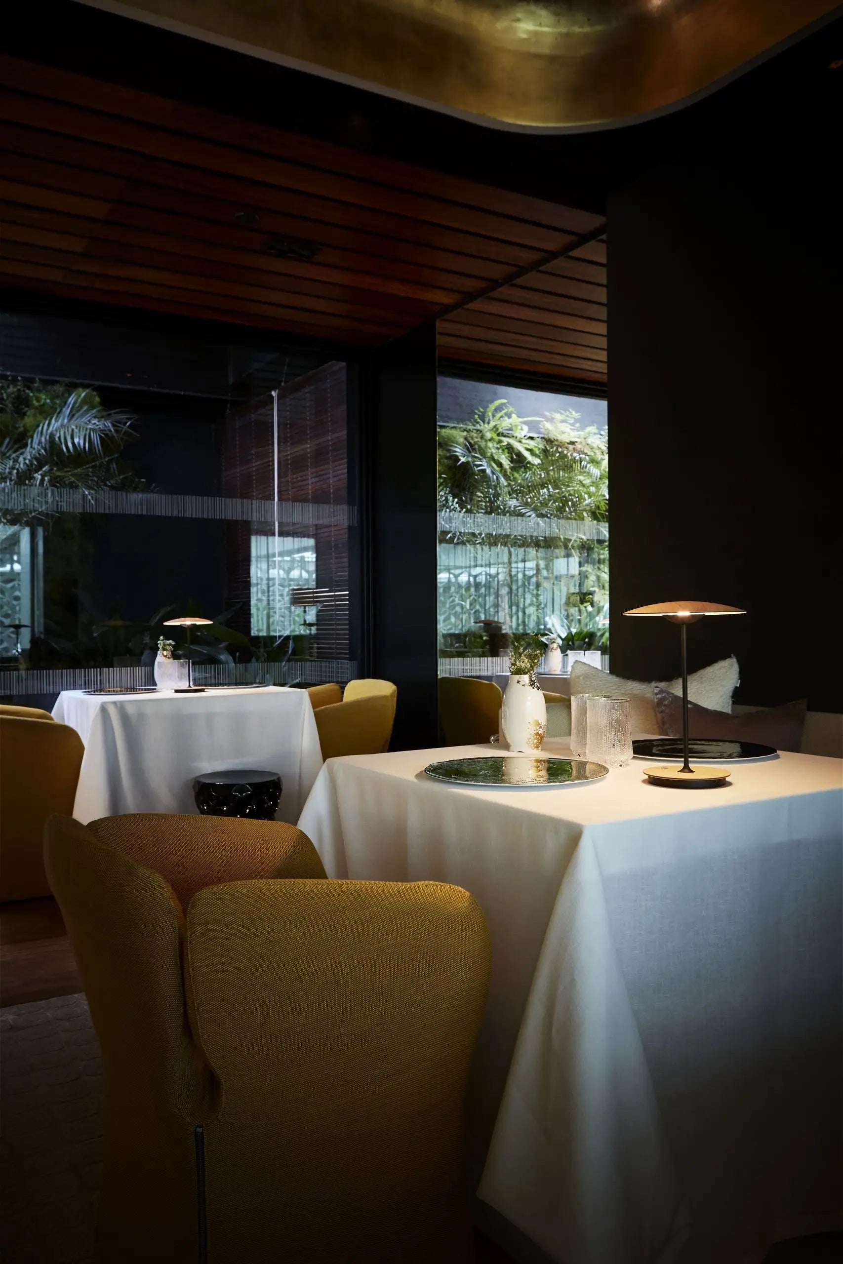 A modern restaurant interior with white tablecloths, soft yellow chairs, Marset Ginger 20 M Table Lamps from Marset casting a gentle glow, and large windows revealing lush greenery outside for an elegant, cosy ambiance.
