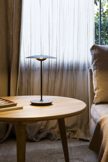 The Marset Ginger 20 M Table Lamp by Marset sits on a round wooden table beside a beige sofa, adding cosy modern style as soft natural light filters through sheer curtains and books rest in the corner.
