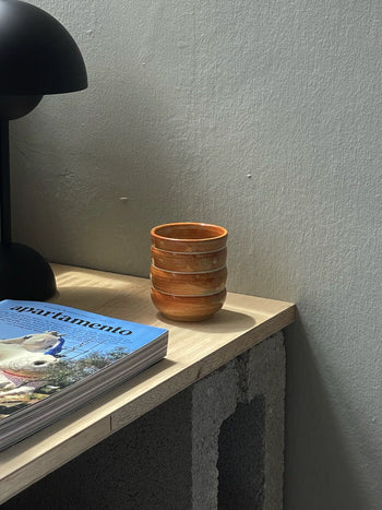 The Mirjam Faber Ceramics Cappuccino Cup Bubble Edition rests on a light wooden table beside a magazine and black lamp, with sunlight casting soft shadows on the textured wall behind.