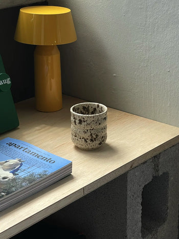 The Mirjam Faber Ceramics Cappuccino Cup Bubble Edition by Mirjam Faber Ceramics rests on a light wooden desk with a concrete base, beside a yellow lamp, green object, and an apartamento magazine with a bird—an elegant Barcelona studio creation.
