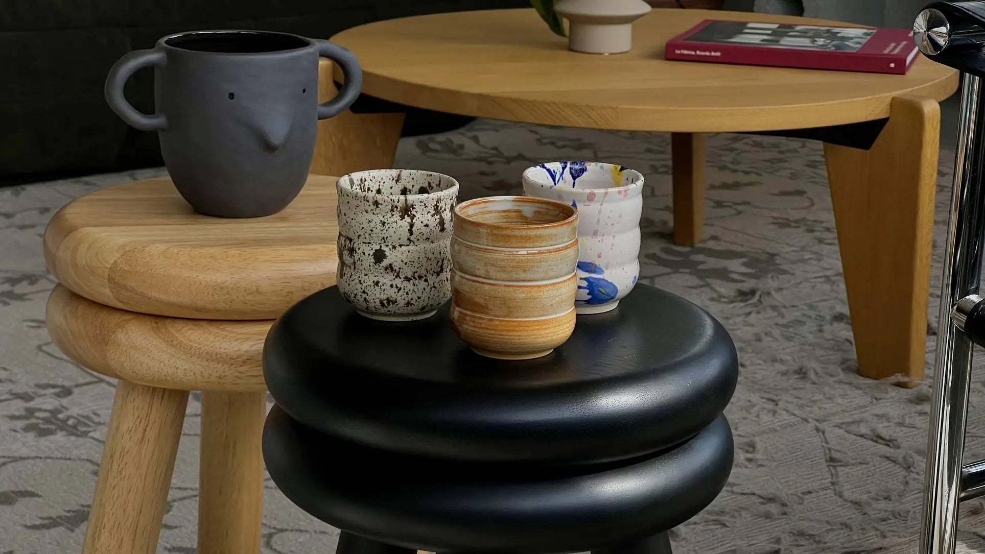 Mirjam Faber Ceramics Cappuccino Cup Bubble Edition cups are displayed on black and wooden stools with a round wooden table and a red book, showcasing the brand’s unique ceramic textures and colors.