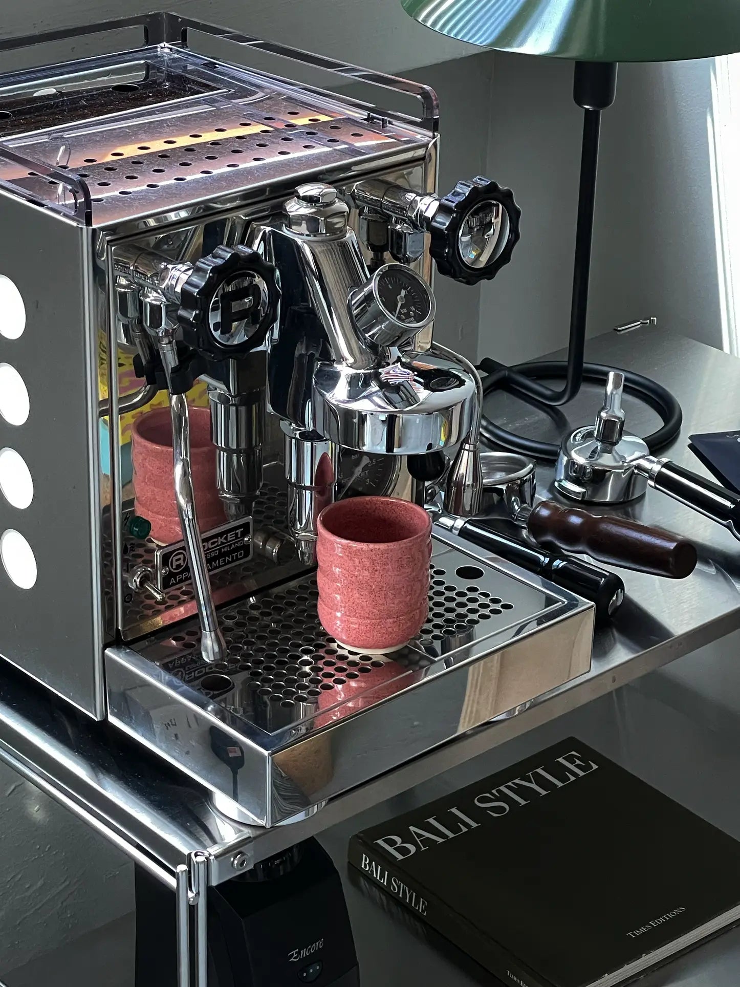 A shiny espresso machine with black dials rests on a metal cart. The Mirjam Faber Ceramics Cappuccino Cup Bubble Edition sits under the spout. Nearby are a tamper, green lamp, phone, and the book BALI STYLE.