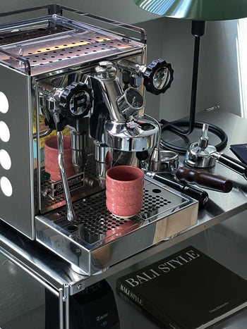 A shiny espresso machine with black dials rests on a metal cart. The Mirjam Faber Ceramics Cappuccino Cup Bubble Edition sits under the spout. Nearby are a tamper, green lamp, phone, and the book BALI STYLE.