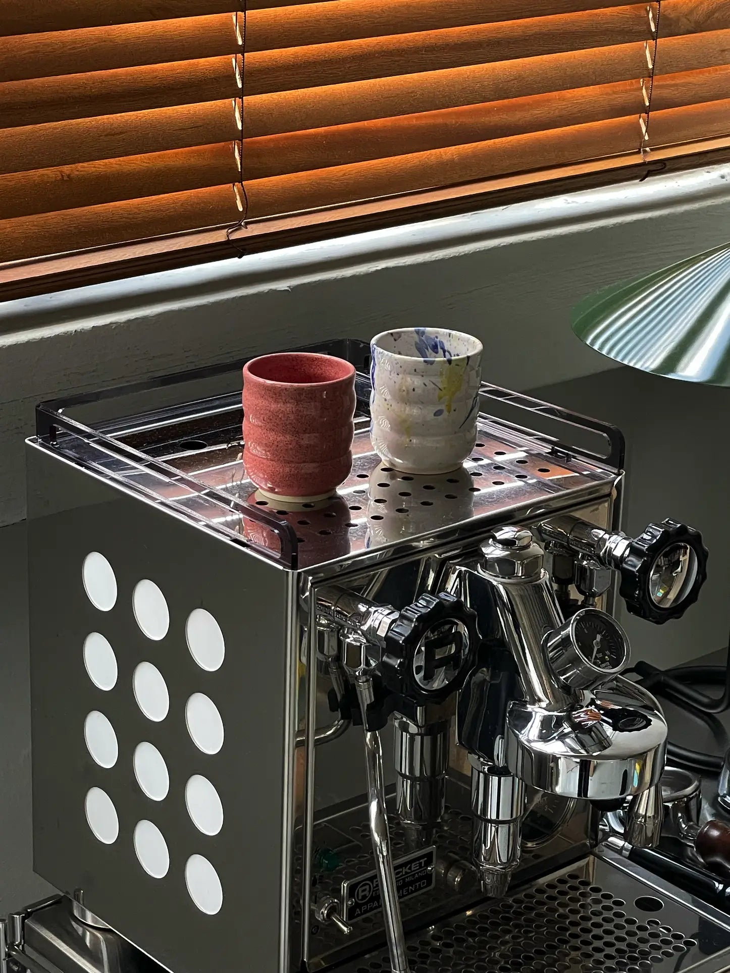 A shiny espresso machine sits topped with two Mirjam Faber Ceramics Cappuccino Cup Bubble Edition cups—one red, one white with blue and yellow bubble spots—by Mirjam Faber Ceramics. Wooden blinds filter sunlight onto the scene.