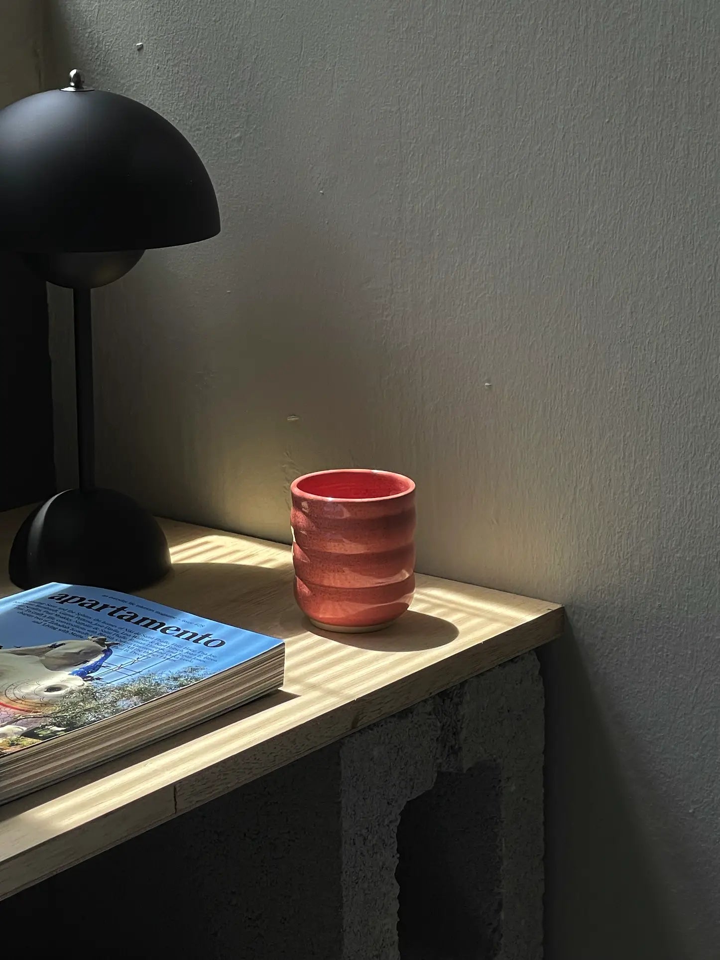 The Mirjam Faber Ceramics Cappuccino Cup Bubble Edition sits on a wooden table beside a lamp and books in soft natural light, casting gentle shadows—a charming touch from Barcelonas Mirjam Faber Ceramics.