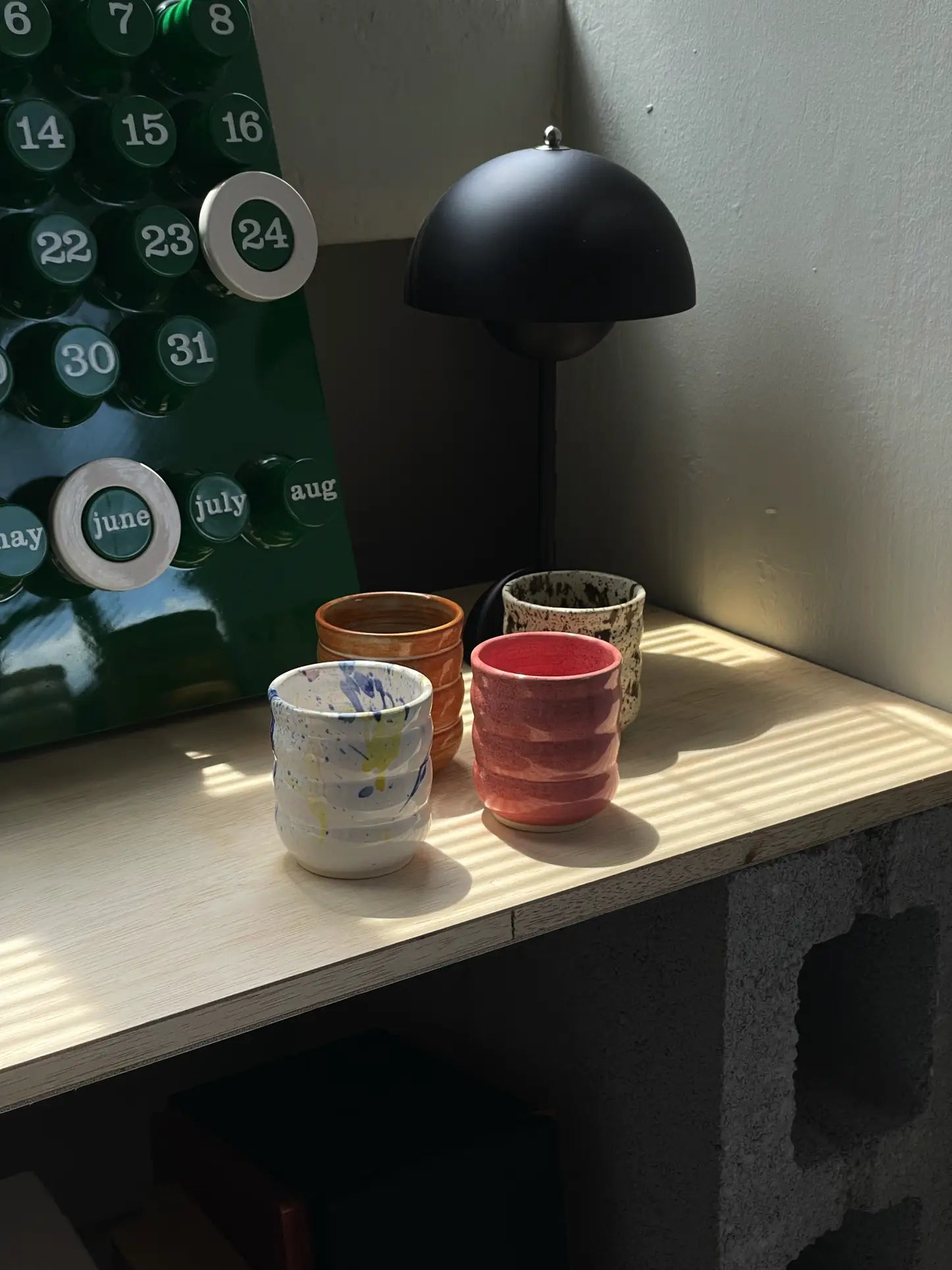 Four vibrant Mirjam Faber Ceramics Cappuccino Cup Bubble Edition cups by Mirjam Faber Ceramics rest on a wooden surface near a green perpetual calendar and black modern lamp, bathed in sunlight from a nearby window.