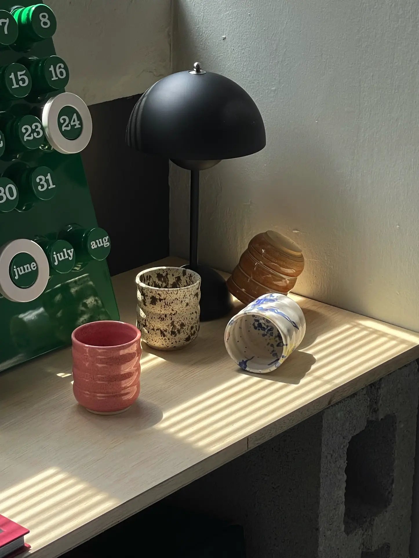 A wooden desk with four Mirjam Faber Ceramics Cappuccino Cup Bubble Edition by Mirjam Faber Ceramics, a black lamp, and a green perpetual calendar open to June and July. Sunlight casts shadows, highlighting the cups and the calendar.