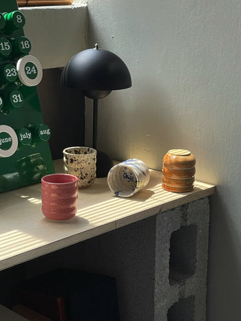 Several Mirjam Faber Ceramics Cappuccino Cup Bubble Edition pieces in various colors and patterns sit on a wooden shelf near a modern black lamp, with sunlight and a green calendar creating a Barcelona ceramics studio vibe.