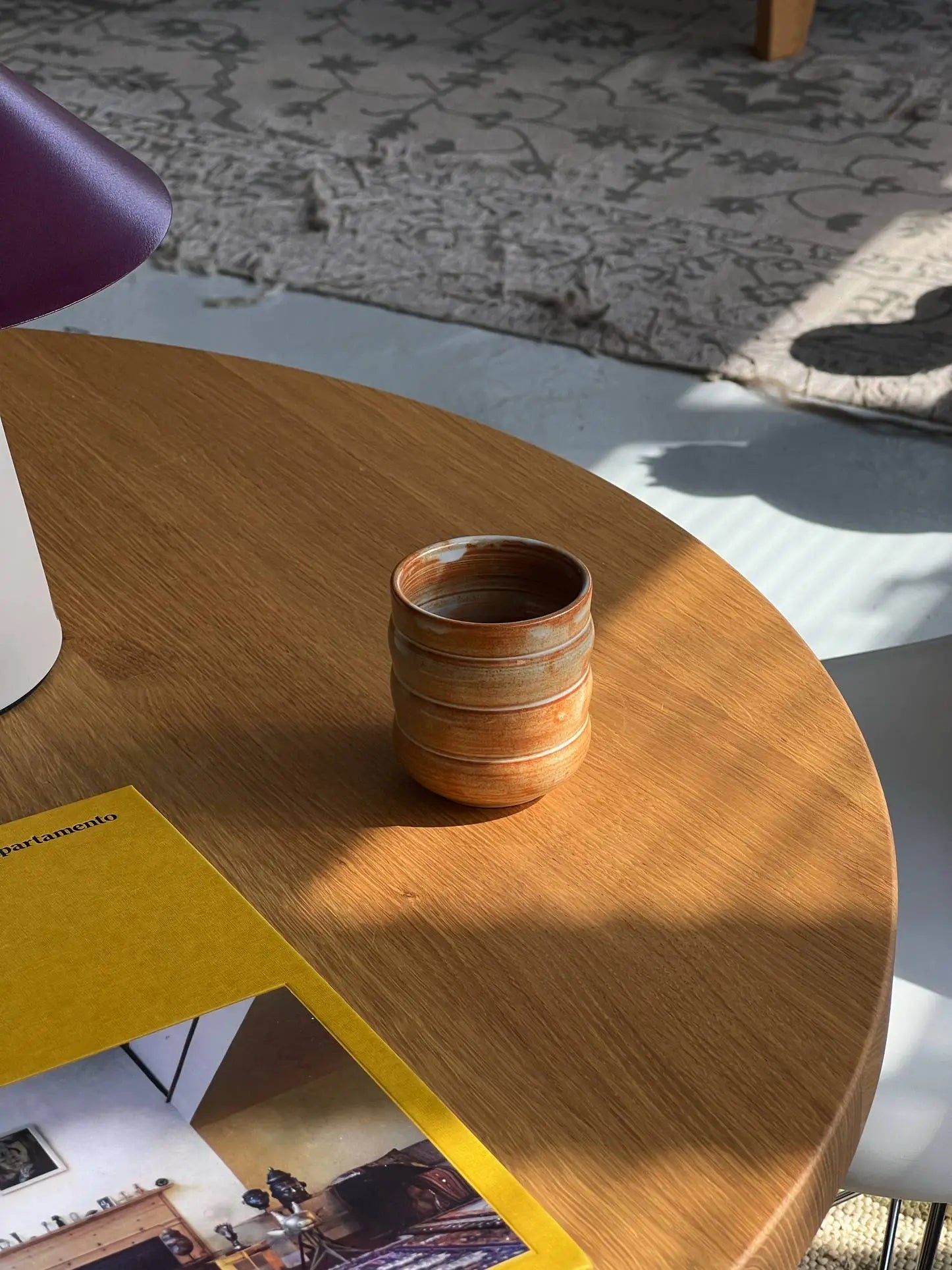 A Mirjam Faber Ceramics Cappuccino Cup Bubble Edition rests on a round wooden table next to a yellow-edged book, sunlight casting shadows. A glimpse of a patterned rug and the brands Barcelona studio artistry appear in the background.