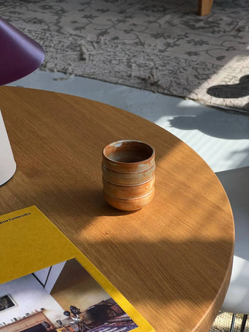 A Mirjam Faber Ceramics Cappuccino Cup Bubble Edition rests on a round wooden table next to a yellow-edged book, sunlight casting shadows. A glimpse of a patterned rug and the brands Barcelona studio artistry appear in the background.