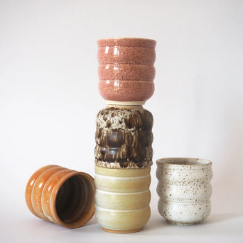 Four Mirjam Faber Ceramics Cappuccino Cup Bubble Edition cups in pink, brown, beige, and speckled white are stacked side by side against a white background—handmade by Mirjam Faber Ceramics in Barcelona.