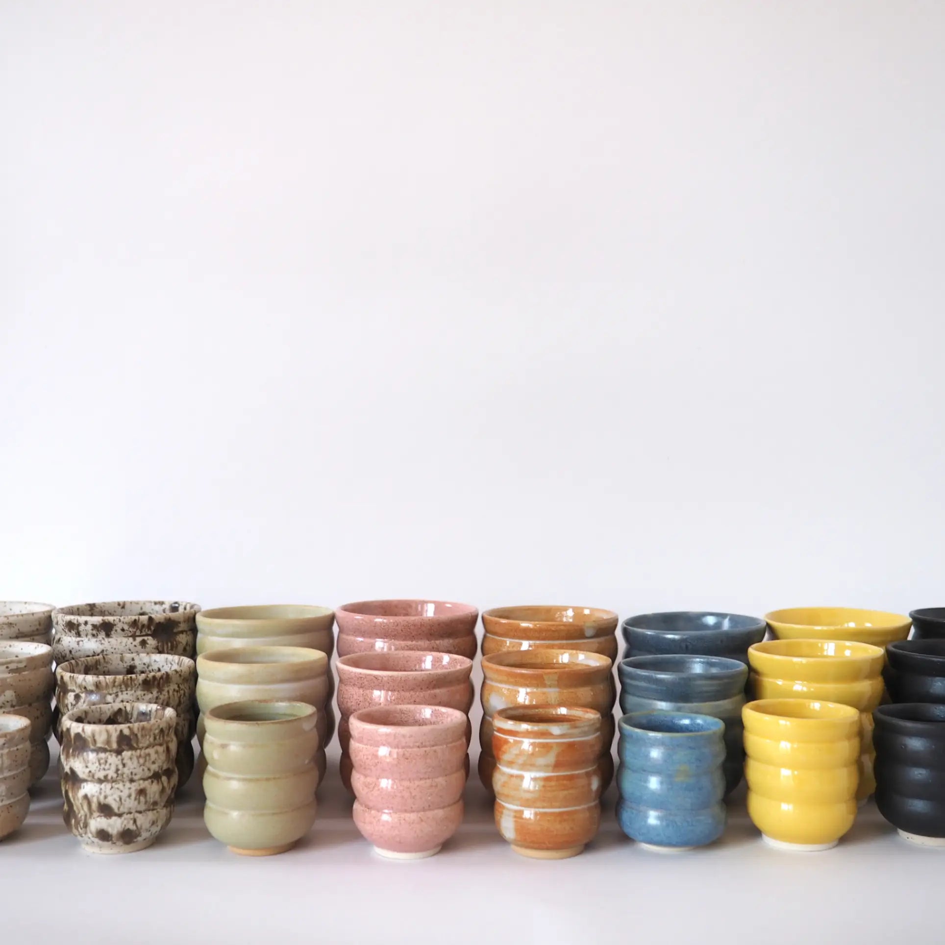 A row of Mirjam Faber Ceramics Cappuccino Cup Bubble Edition in speckled, light green, pink, orange, blue, yellow, and black glazes is neatly arranged against a plain white background from Mirjam Faber Ceramics Barcelona studio.