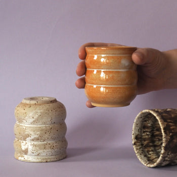 A hand holds a small, round, orange Mirjam Faber Ceramics Cappuccino Cup Bubble Edition with horizontal ridges. Two more cups by Mirjam Faber Ceramics—one speckled beige, one brown—are nearby against a light purple background.