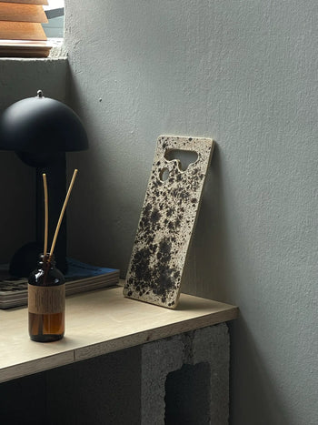 A speckled phone case rests on a wooden desk near a reed diffuser, black lamp, and magazines, evoking the rustic texture and unique cutouts of the Mirjam Faber Ceramics Sushi Plate by Mirjam Faber Ceramics.