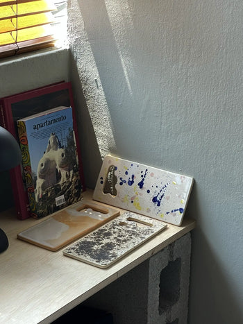 A small desk holds three phone cases—paint splatter, clear, and brown-patterned—next to a book titled apartamento, a black lamp, and Mirjam Faber Ceramics Sushi Plates by Mirjam Faber Ceramics, all near a sunlit window.